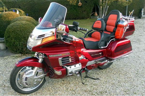 Rotes Tourenmotorrad Honda Gold Wing mit Chromdetails und geräumiger Sitzfläche, geparkt auf einem Kiesweg umgeben von Grün.